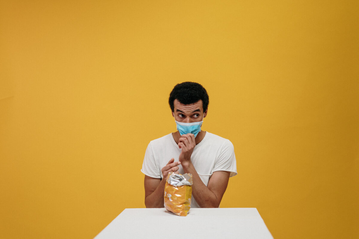 Man met mondkapje voor een gele muur met een zak chips
