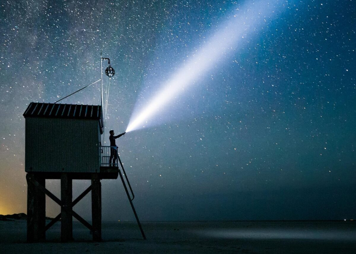 Man die met zaklamp in een donkere zee schijnt vanaf een strandwachthuisje onder een sterrenhemel in Friesland - Photo Hans Jellema en Holland Boven Amsterdam